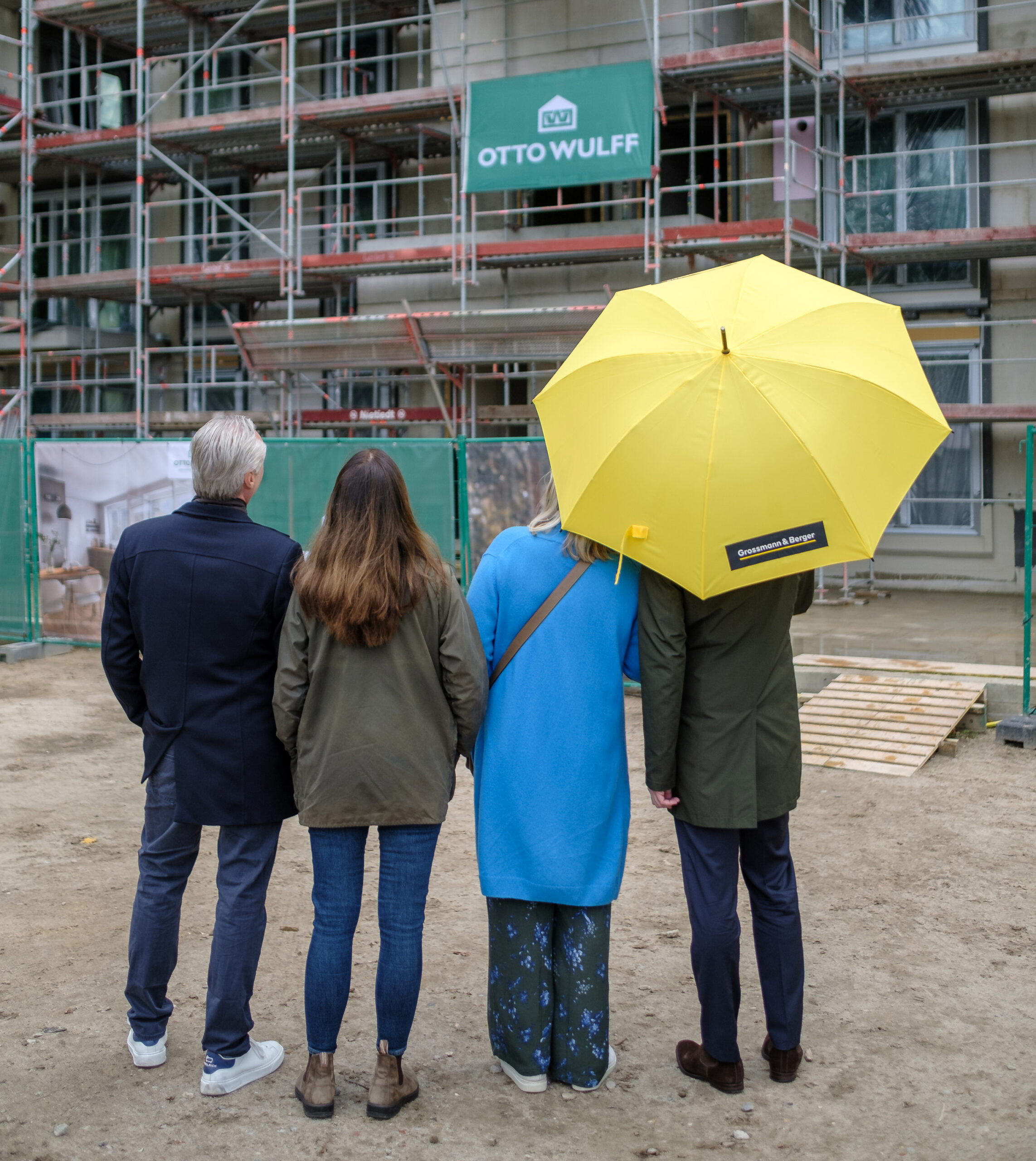 Vier Personen stehen vor einer Baustelle, einer hält einen gelben Regenschirm.