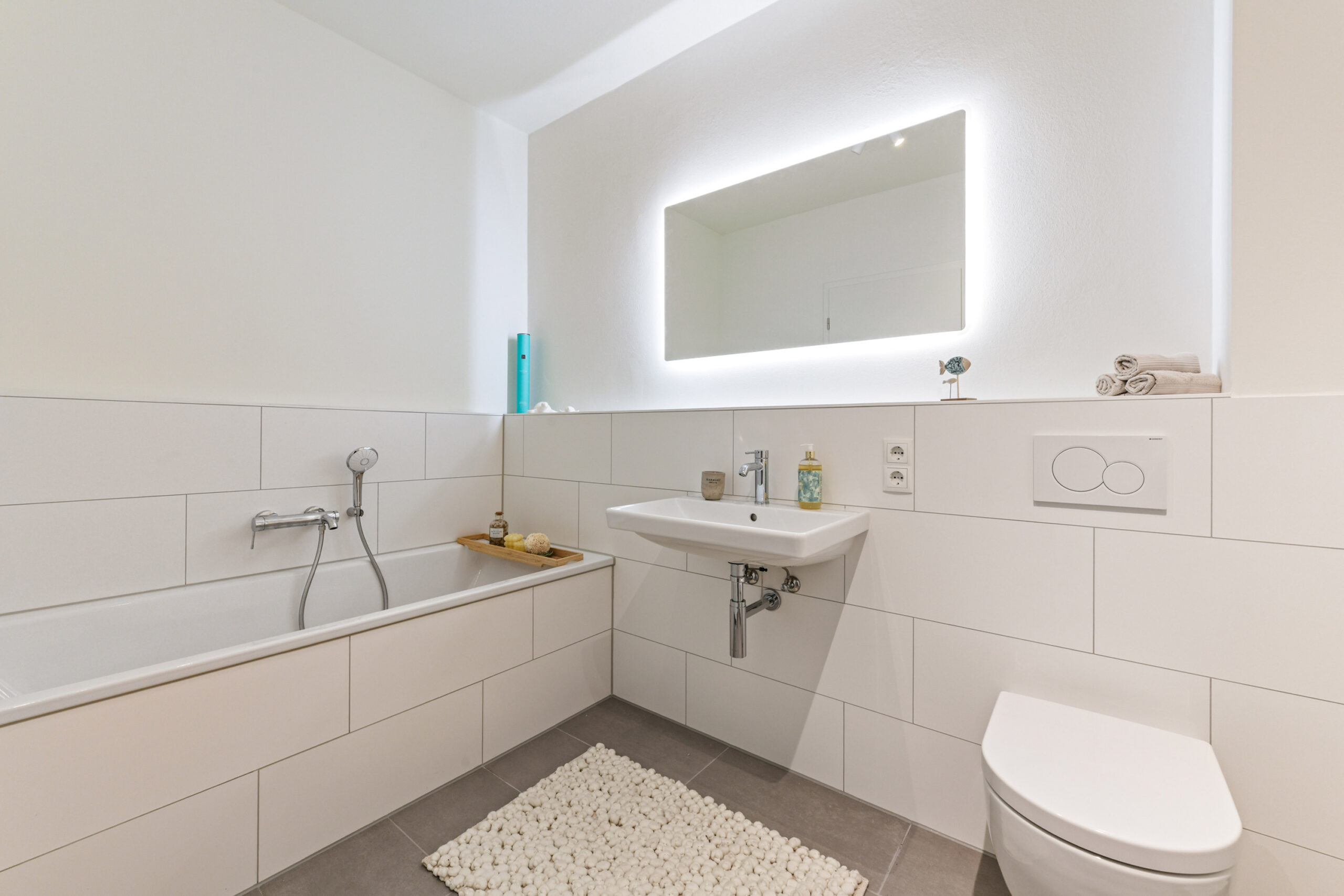 Modernes, weiß gefliestes Bad mit Badewanne, Waschbecken und beleuchtetem Spiegel.